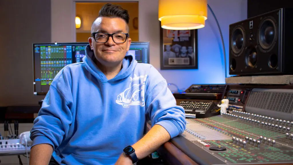 Dana Nielsen in his recording studio, seated, facing camera, smiling, wearing blue hoodie, resting elbow on mixing console