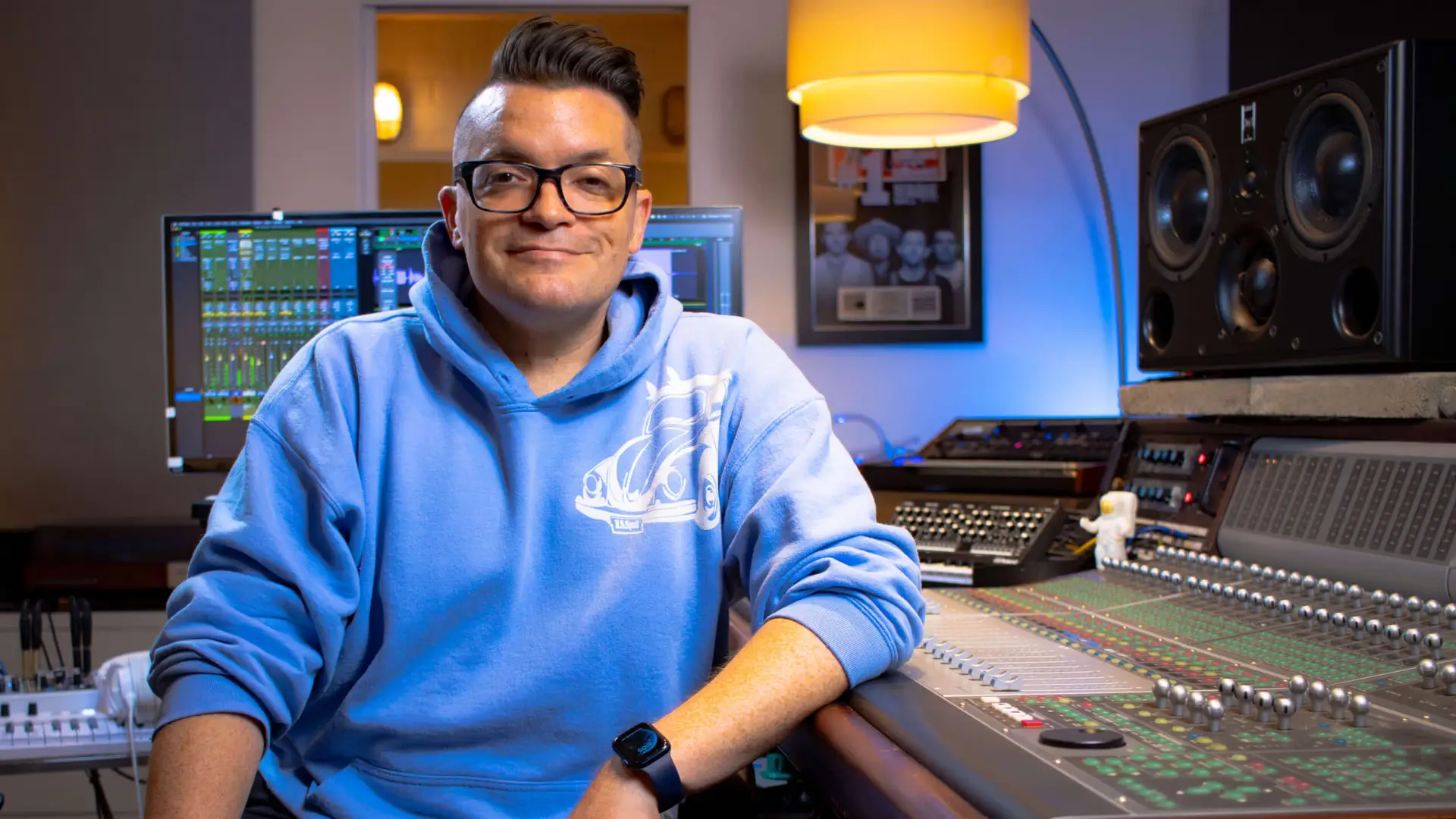 Dana Nielsen in his recording studio, seated, facing camera, smiling, wearing blue hoodie, resting elbow on mixing console