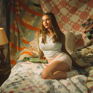Singer-songwriter, Cat Matthews, seated on bed with quilts. Cover art from her album single, "Greener Pastures / Happiness Is Sad"